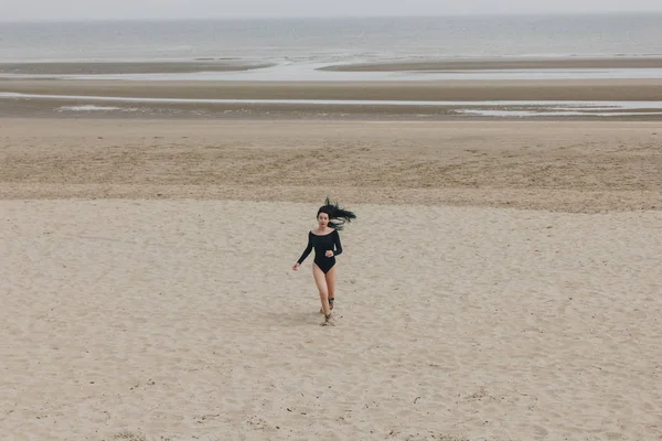 Attractive young woman in black bodysuit running on sandy seashore — Stock Photo