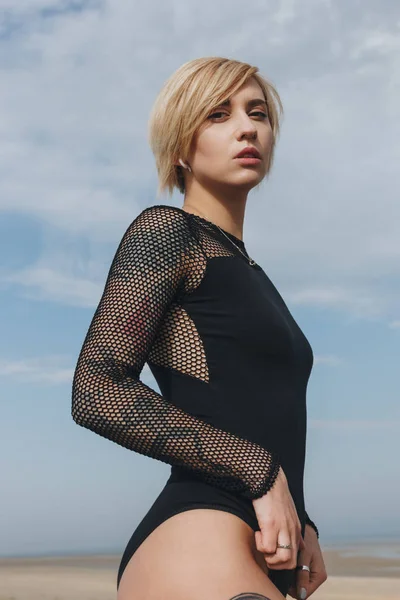 Vista inferior de sexy jovem mulher em preto bodysuit no céu nublado fundo — Fotografia de Stock