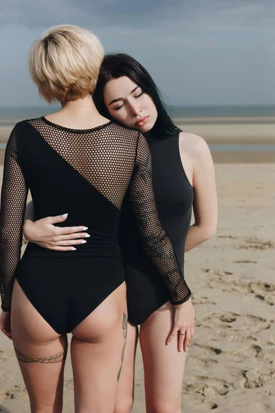 Mujeres jóvenes atractivas en trajes negros abrazándose en la playa de arena en el día nublado - foto de stock