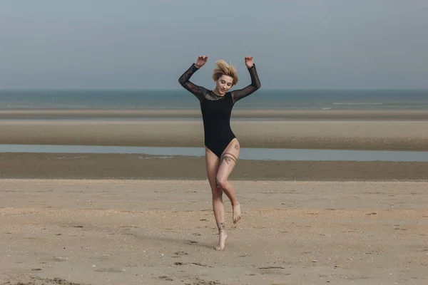Bella giovane donna in body nero saltando sulla riva nella giornata nuvolosa — Foto stock