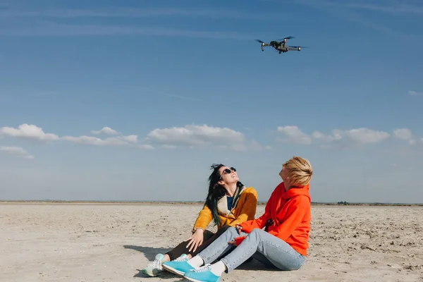 Stylish girls sitting on beach and looking at drone, Saint michaels mount, Normandy, France — Stock Photo