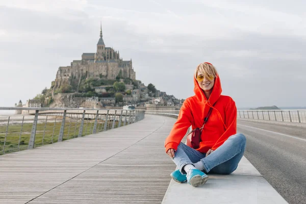 Feliz joven mujer sonriendo a la cámara mientras se sienta cerca de mont saint michel, francia - foto de stock