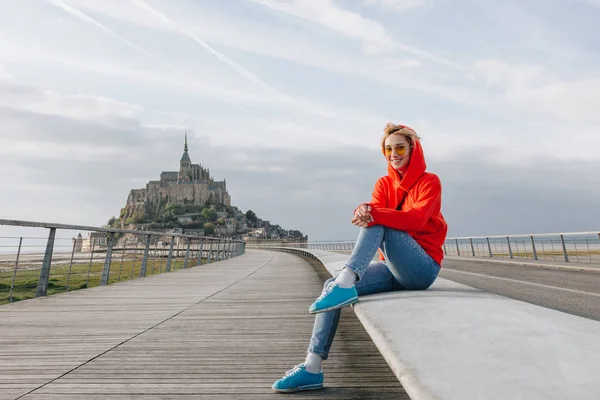 Beautiful happy girl in sunglasses smiling at camera while sitting near mont saint michel, france — Stock Photo