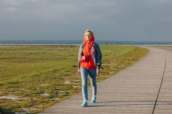 Vista completa di bella giovane donna in occhiali da sole che cammina su passerella in legno sulla costa del mare — Foto stock