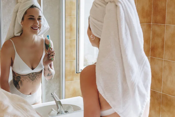 Красива усміхнена молода жінка в нижній білизні тримає зубну щітку і дивиться на дзеркало у ванній — стокове фото