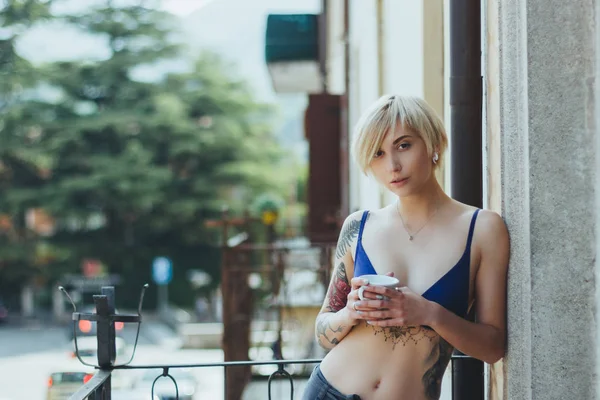 Hermosa mujer joven en sujetador sosteniendo taza y mirando a la cámara mientras está de pie en el balcón - foto de stock