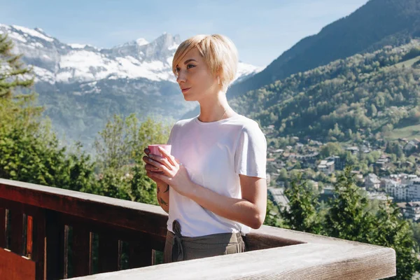 Young woman holding cup and looking away while standing on balcony with beautiful mountains behind, mont blanc, alps — Stock Photo