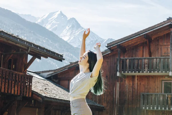 Side view of beautiful young woman raising hands while standing between wooden houses in mountain village, mont blanc, alps — Stock Photo