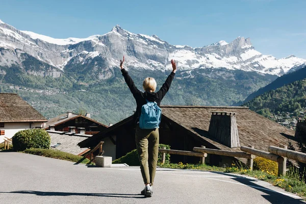 Back view of girl with backpack raising hands and walking in scenic mountain village, mont blanc, alps — Stock Photo