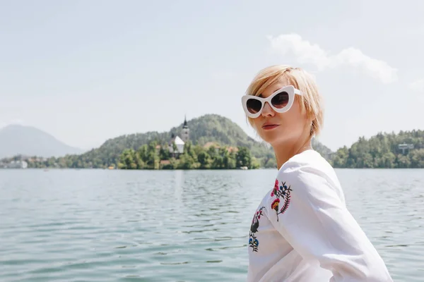 Hermosa mujer joven en gafas de sol de pie cerca de lago escénico en las montañas, sangró, slovenia - foto de stock