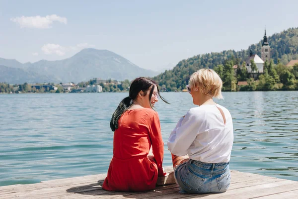 Back view of stylish girls sitting on wooden pier near tranquil mountain lake, bled, slovenia — Stock Photo
