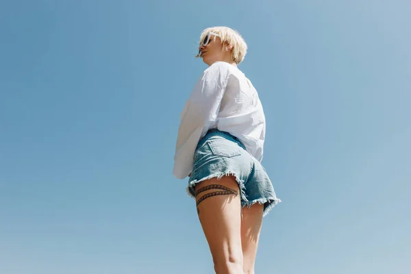 Низкий угол зрения на красивую молодую стильную женщину в солнцезащитных очках и джинсовых шортах, стоящих против ясного голубого неба — стоковое фото