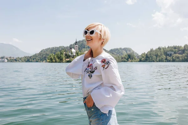 Belle jeune femme souriante dans des lunettes de soleil debout près du majestueux lac de montagne, saigné, slovenia — Photo de stock