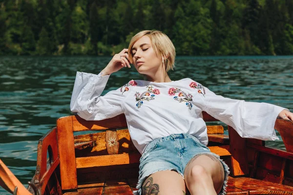 Beautiful young woman with closed eyes sitting in wooden boat on lake — Stock Photo