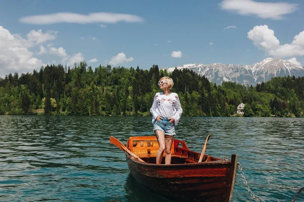 Beautiful young woman in sunglasses standing in boat at tranquil mountain lake, bled, slovenia — Stock Photo