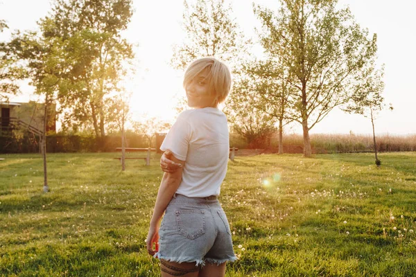 Beautiful young woman in t-shirt and denim shorts looking at camera while standing on green lawn at sunset — Stock Photo