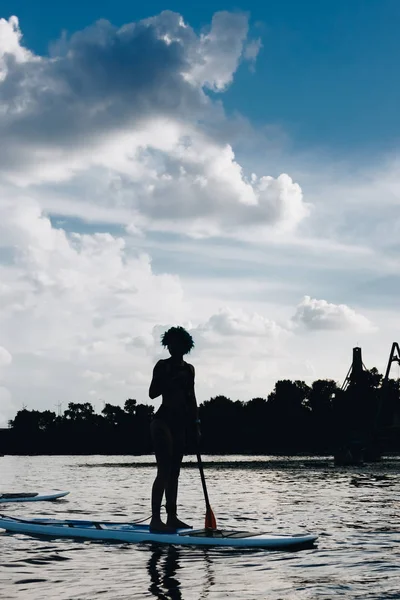 Silhouette de sportive debout sur une planche à pagaie sur une rivière avec un ciel nuageux — Photo de stock
