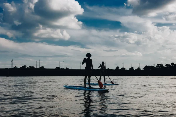 Silhouettes of athletic women paddle boarding on river with cloudy sky — Stock Photo
