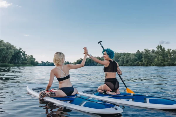 Sportliche Frauen geben Highfive, während sie auf Paddelbrettern auf dem Fluss sitzen — Stockfoto