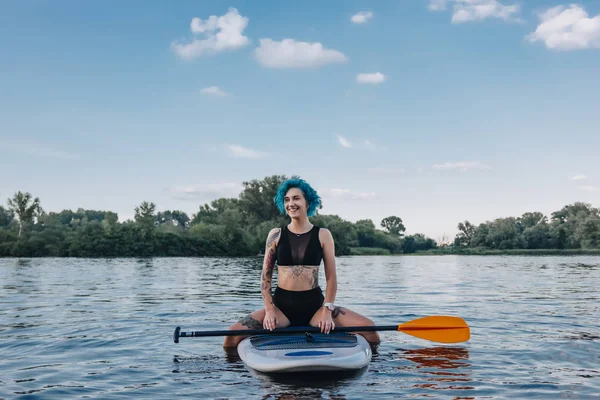 Beautiful tattooed girl with blue hair sitting on sup board on river — Stock Photo