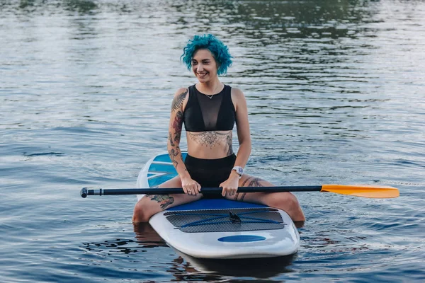 Lächelndes Mädchen mit blauen Haaren sitzt auf einem Paddelbrett auf dem Fluss — Stockfoto