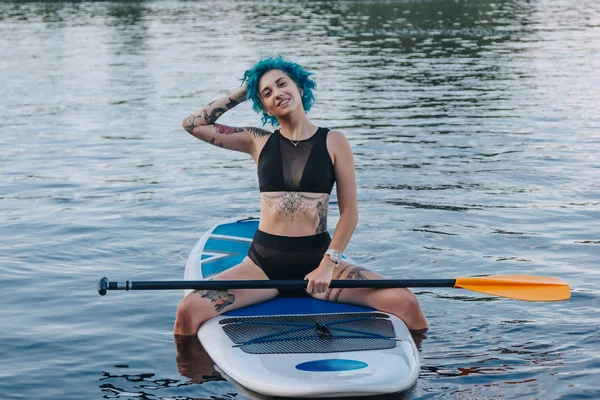 Sorridente ragazza tatuata con i capelli blu appoggiata sul paddle board sul fiume — Foto stock