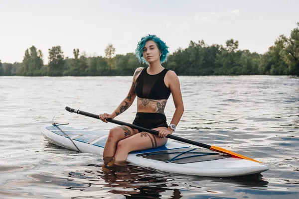 Menina tatuada atraente com cabelo azul descansando em paddleboard no rio — Fotografia de Stock