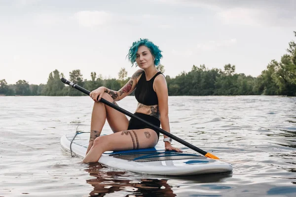 Beautiful tattooed girl with blue hair sitting on paddleboard on river — Stock Photo