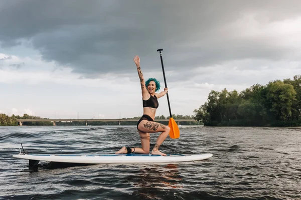 Aufgeregtes, sportlich tätowiertes Mädchen mit blauen Haaren winkt und paddelt auf dem Fluss — Stockfoto