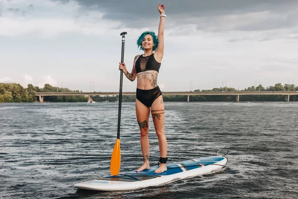Tattooed sportswoman waving and paddle boarding on river in summer — Stock Photo