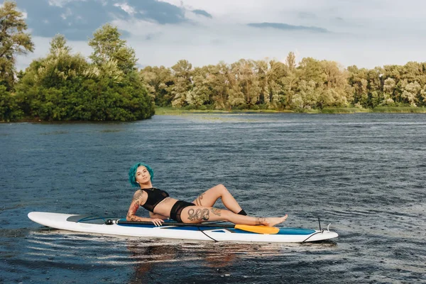 Привлекательная татуированная девушка с голубыми волосами расслабляется на веслах на реке — стоковое фото