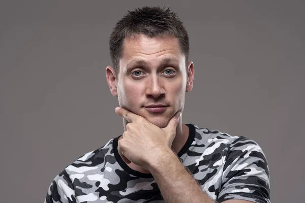 Portrait of expressive uncertain man looking at camera with hands on chin over gray background.