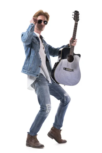 Confident young redhead man in jeans posing with acoustic guitar holding sunglasses. Full body isolated on white background.