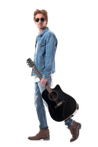 サングラスを歩くと後ろを見てギターを運ぶジーンズでクールな自信を持った男のプロフィールビュー 白地に隔離された全身 ストック画像
