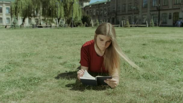 Vacker flicka läser en bok på campus gräsmatta — Stockvideo