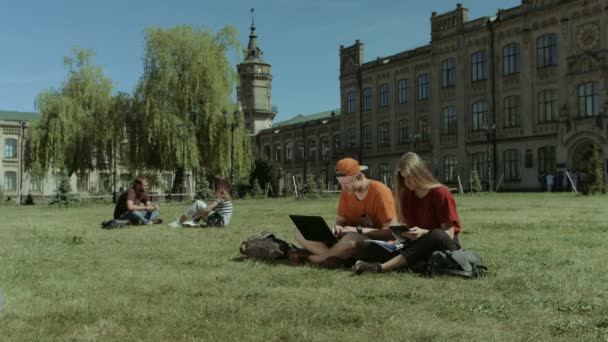 Estudiantes con dispositivos electrónicos que estudian en el césped — Vídeo de stock