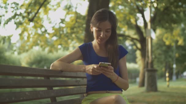 Улыбающаяся красотка пишет смс-ки на сотовый в парке — стоковое видео
