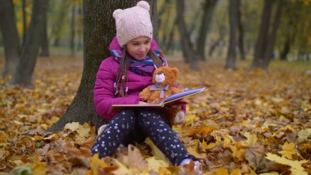 Милая девушка читает книгу под елкой в осеннем парке — стоковое видео