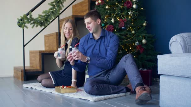 恋冬の休日にクリスマス ツリーの下に座っている間石工の Jar ファイルからホット チョコレート飲み物を楽しむ陽気な素敵なカップル 新しい年を祝うとすすりながら肯定的なカップル ホット ドリンク自宅 — ストック動画