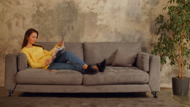 Прекрасна дівчина читає журнал і розслабляється на дивані — стокове відео