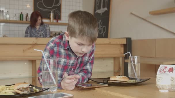 Niño pequeño jugando en el teléfono mientras come en la cafetería — Vídeo de stock