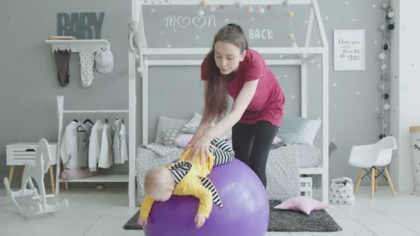 Cheerful mom training baby girl on fitball indoor — Stock Video