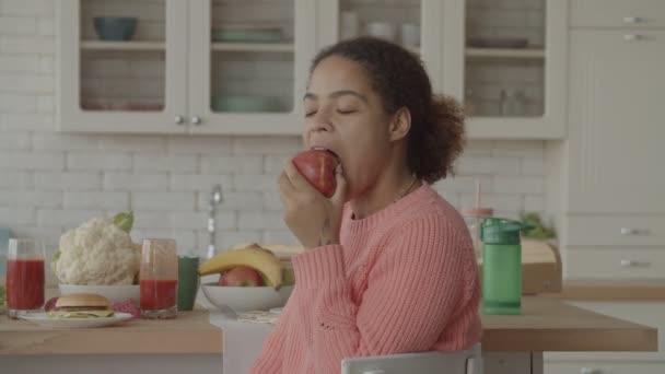 Mujer africana sonriente comiendo manzana jugosa en la cocina — Vídeos de Stock