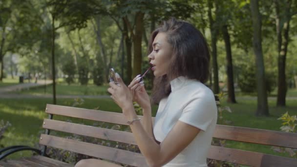 Preciosa hembra negra aplicando lápiz labial en el parque — Vídeo de stock