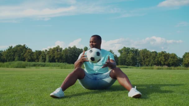 Позитивный человек с футбольным мячом сидит на зеленом поле — стоковое видео