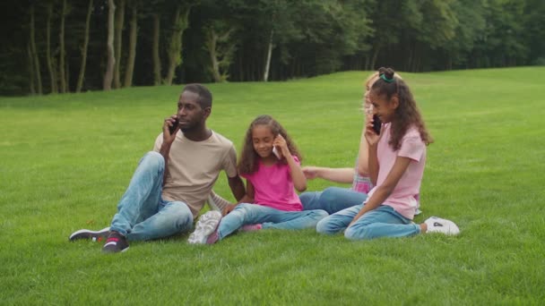 Багатоетнічна сім'я зайнята смартфонами на відкритому повітрі — стокове відео