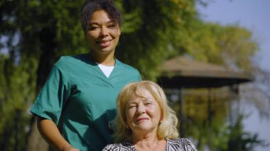 Mutlu, emekli, güzel, yaşlı kadın tekerlekli sandalye kullanıcısı ve üniformalı, dost canlısı siyahi kadın portresi. Olumlu ifadelerle bakıyor, yaz doğasında anlayış ve birlik gösteriyor..