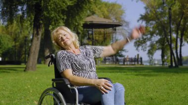 Tekerlekli sandalyedeki olumlu yaşlı kadın yaz doğasında rehabilitasyon sırasında egzersiz yapıyor, kaslarını esnetiyor ve güçlendiriyor..