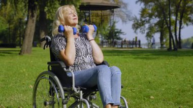 Fiziksel engelli, çekici, yaşlı kadın tekerlekli sandalye kullanıcısı. Halter pazıları kıvrılıyor, emekliliği ve boş zamanlarını halk parkında geçirirken kol kaslarını güçlendiriyor..