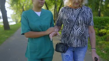 Üniformalı Afrikalı Amerikalı kadın bakıcı, yaz parkında yürüyen bastonlu, yürüyen ve eğlenen yaşlı bir kadının elini tutuyor ve destekliyor..